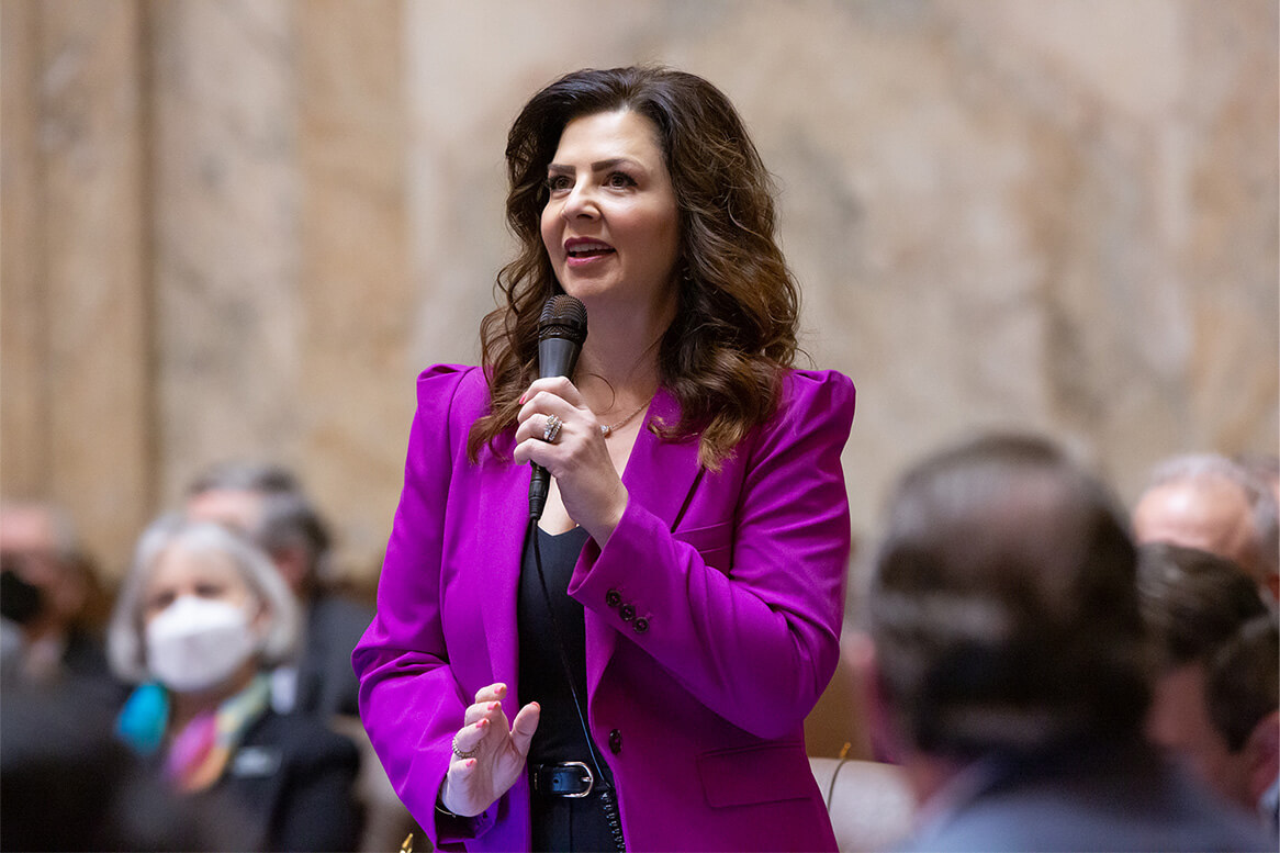 Kelly Chambers in purple suit speaking into a microphone in a crowded room